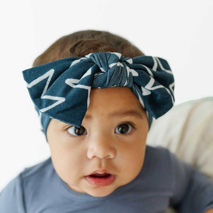 A baby wearing a dark teal blue fabric bow headband with a white star pattern, paired with a slate blue long-sleeve shirt, photographed against a bright white background.