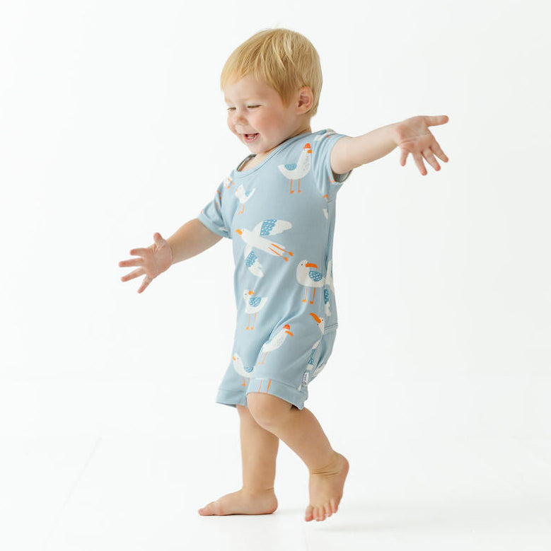 A toddler with blonde hair wearing a light blue short-sleeve romper with a white and orange bird pattern, walking barefoot on a white studio background.