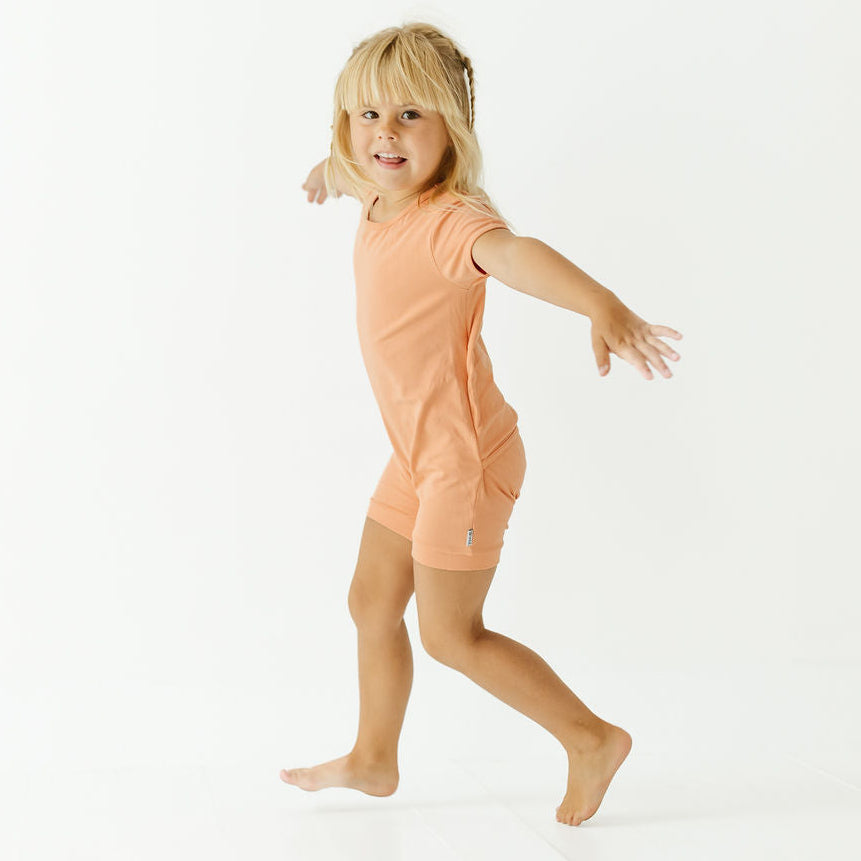 A young blonde girl wearing a peach-colored short-sleeve t-shirt and matching shorts, captured mid-step in a playful pose against a seamless white studio background.