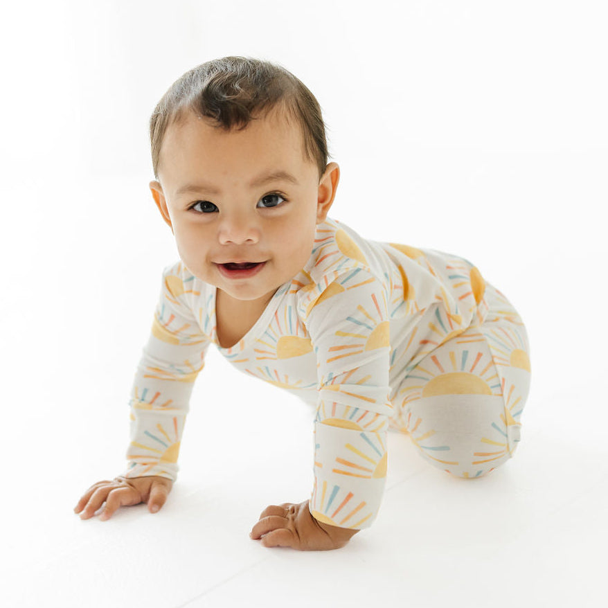 A baby crawling on a white studio floor wearing a long-sleeve one-piece pajama with a yellow and orange sunburst pattern on a white background.