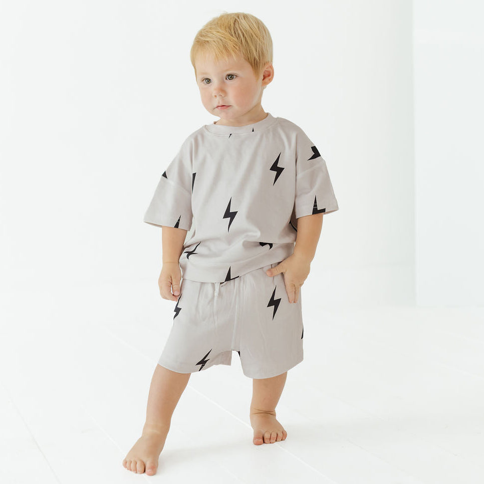 A toddler standing barefoot in a bright white studio, wearing a light gray short-sleeve t-shirt and matching shorts with a black lightning-bolt print.