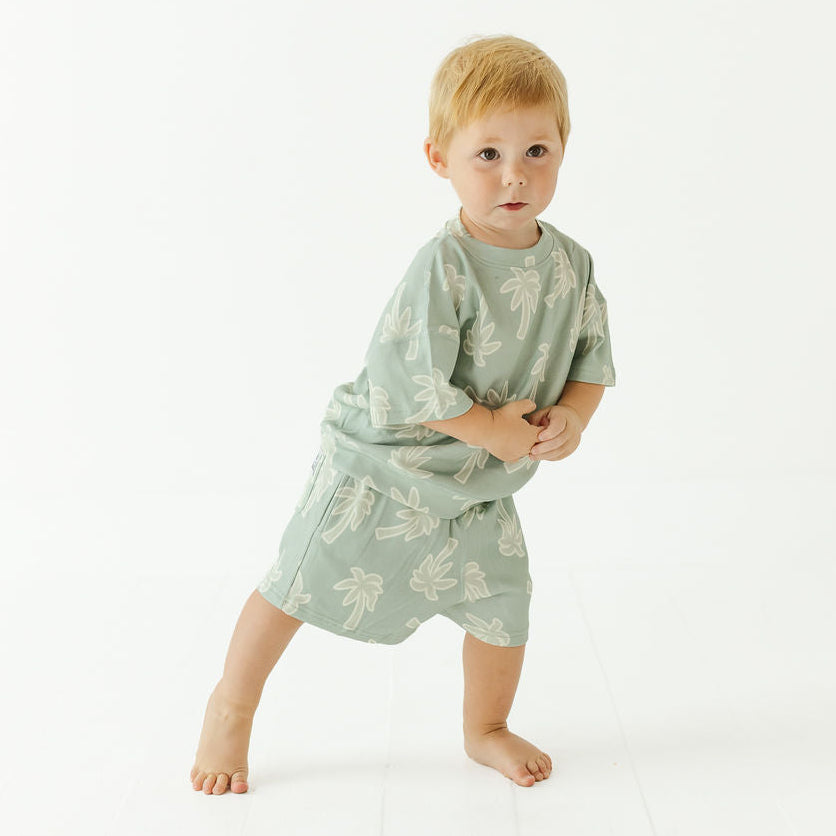 A toddler standing barefoot in a studio, wearing a light sage green short-sleeve pajama set with a white leaf pattern.