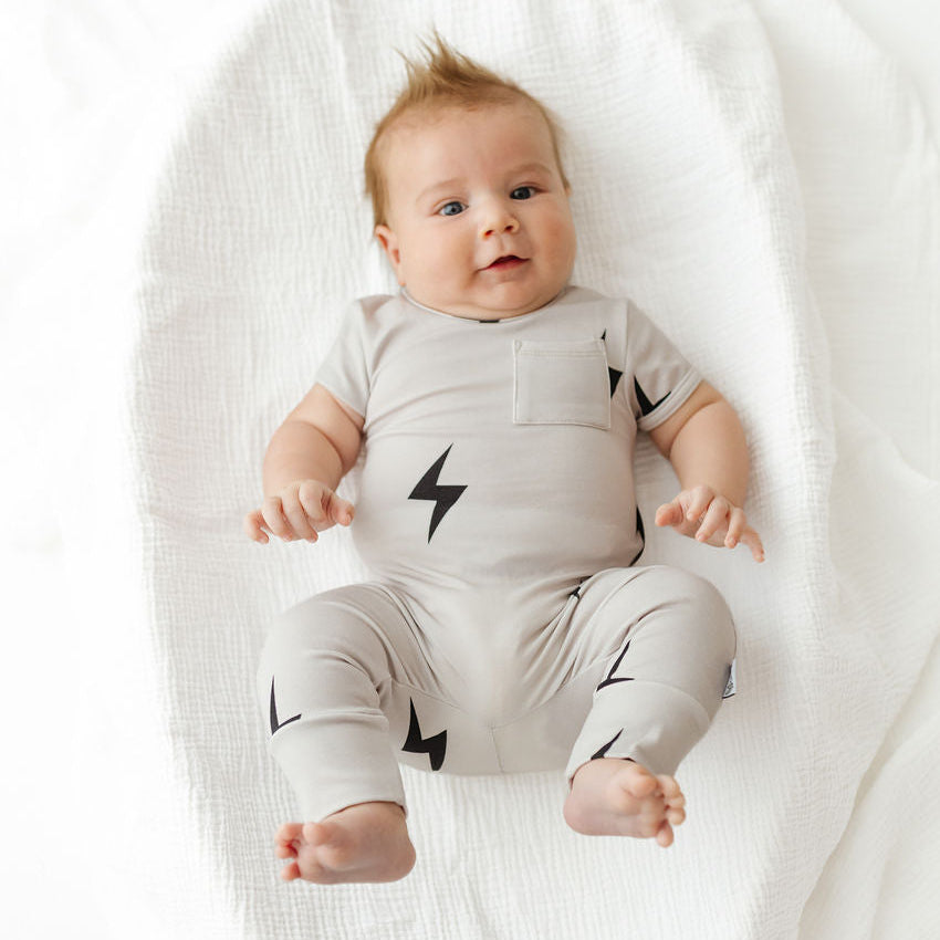 A baby lying on a white muslin blanket, wearing a light gray short-sleeve romper and matching leg warmers with black lightning bolt prints.