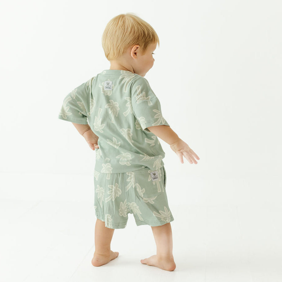 A toddler standing barefoot on a white studio background, viewed from behind, wearing a light sage green short-sleeve pajama set with a white botanical leaf pattern.