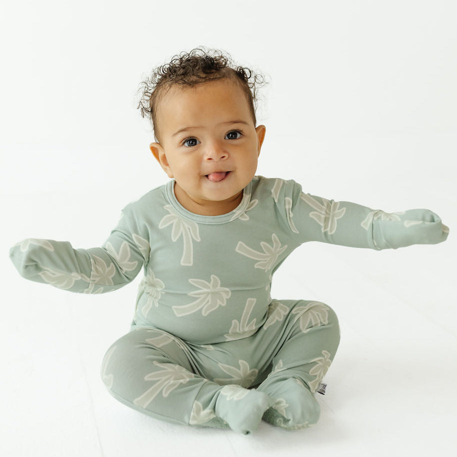 A baby sitting cross-legged wearing a sage green footed sleeper with a white palm tree pattern, photographed against a clean white background.