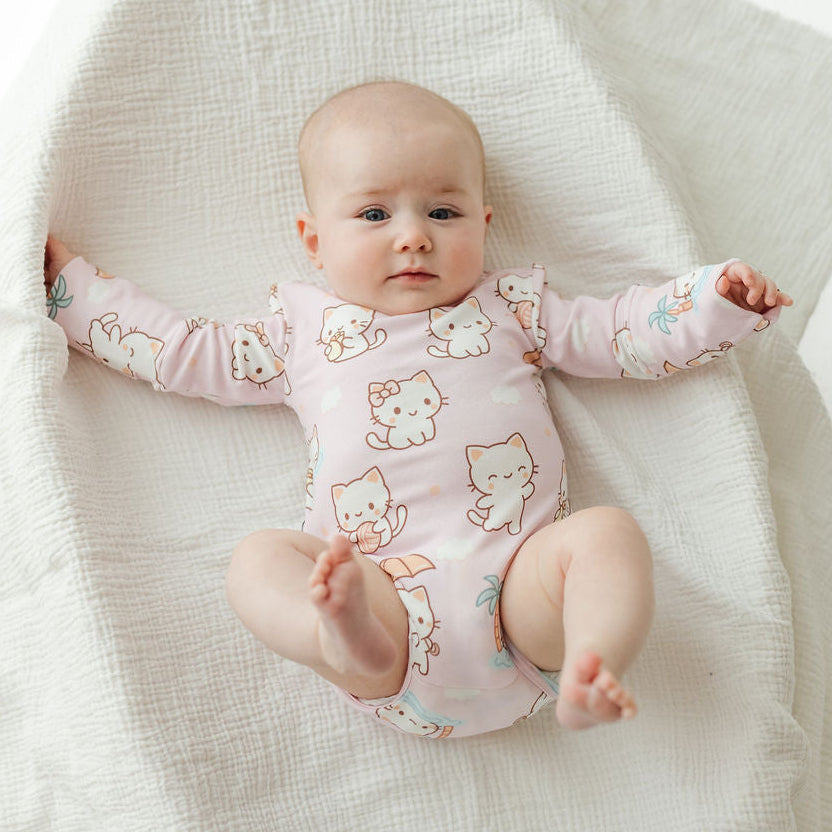 A baby lying on a textured white muslin blanket, wearing a light pink long-sleeve bodysuit with a cute cartoon cat print.