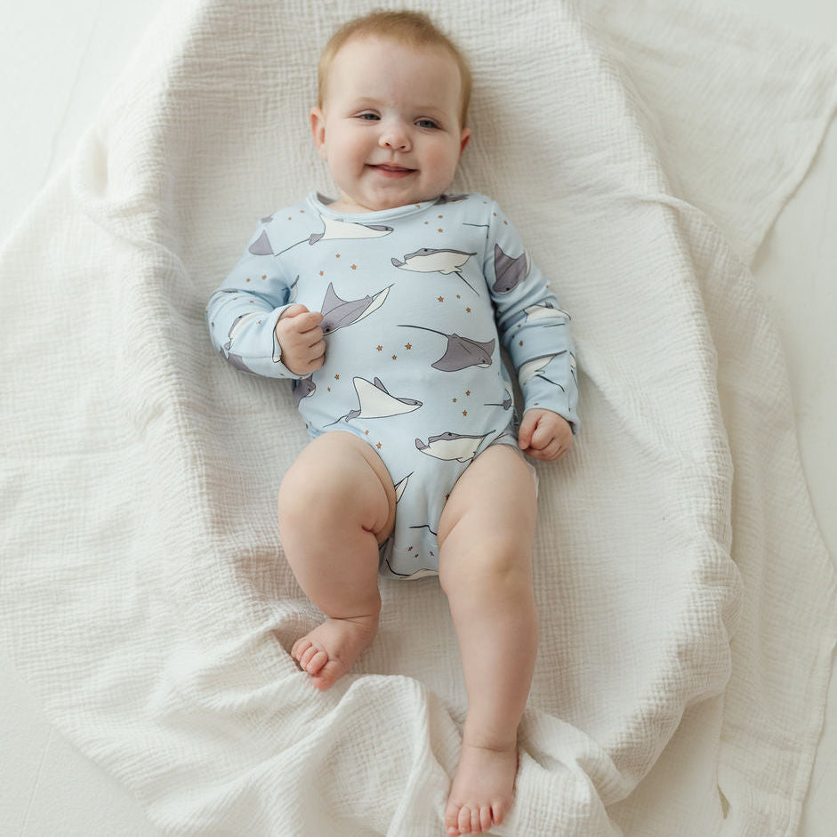 A baby lying on a white textured muslin blanket, wearing a light blue long-sleeve bodysuit with a gray and white whale print.