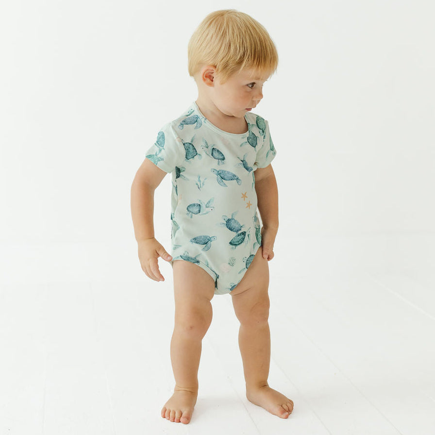 A toddler standing barefoot on a white studio floor, wearing a short-sleeve light green bodysuit with a blue and green fish print.