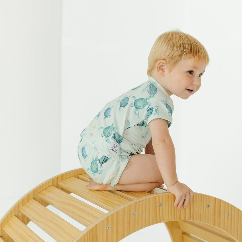 A toddler with blonde hair sits on a curved wooden play structure, wearing a short-sleeve light green romper with a blue-green sea turtle pattern. The photo is taken in a bright, minimalist room with white walls.