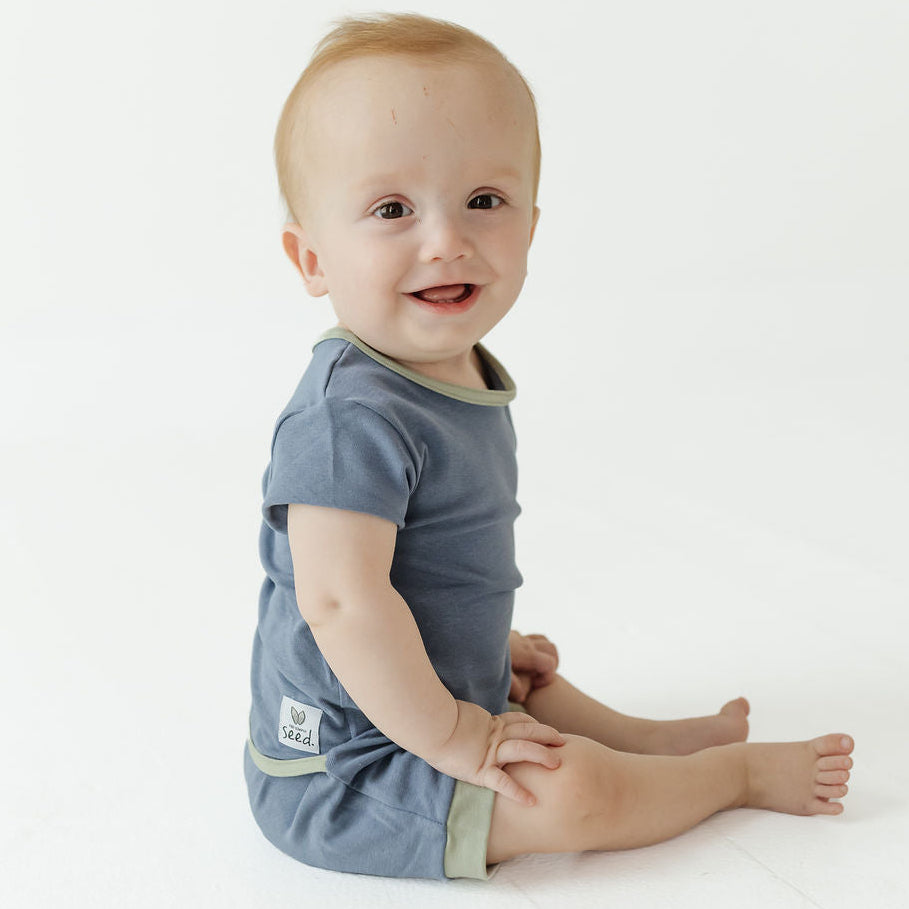 A baby sitting on a white studio floor wearing a slate blue short-sleeve romper with sage green trim.