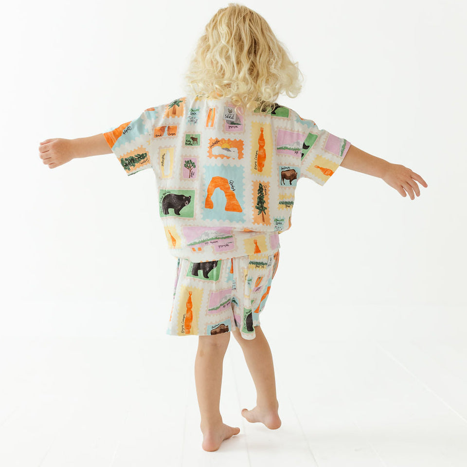 A child with curly blonde hair is shown from behind wearing a matching short-sleeve robe and shorts set with a colorful landscape patchwork print, set against a plain white studio background.