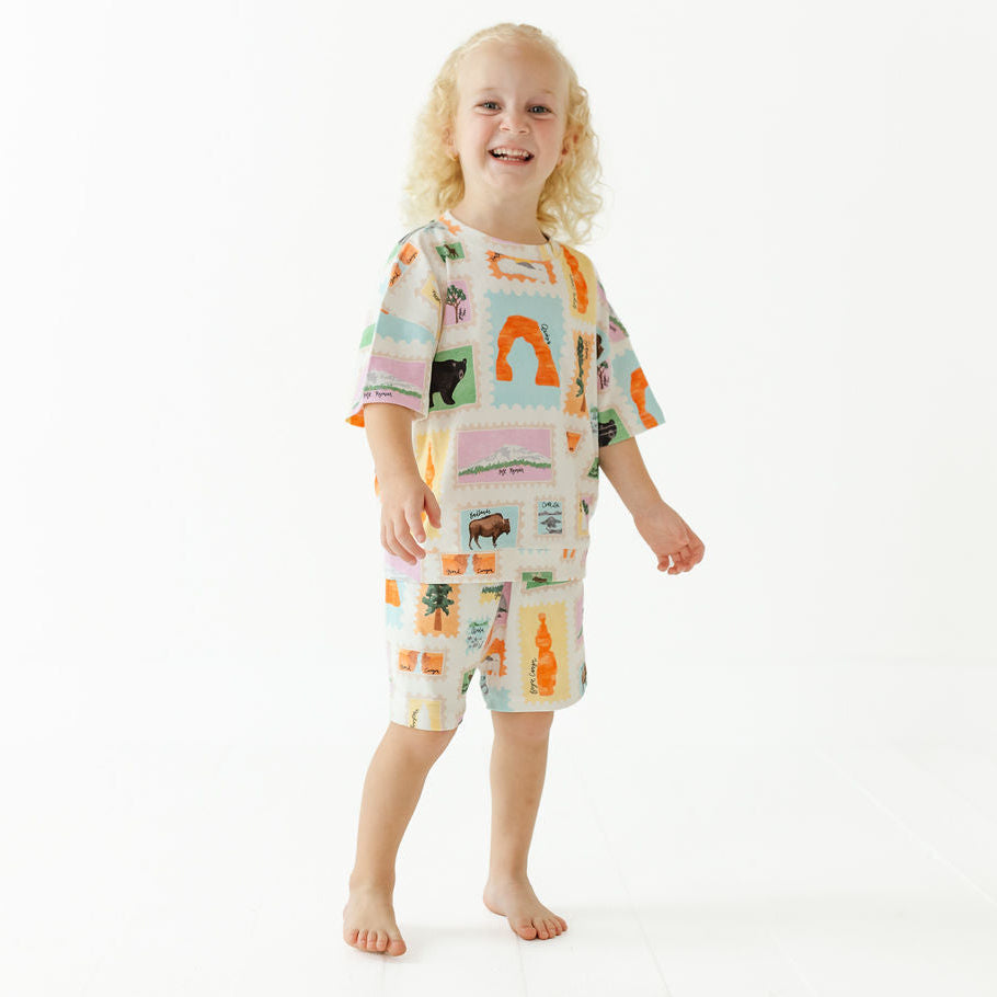 A young child with curly blonde hair stands barefoot against a plain white studio background, wearing a matching short-sleeve pajama set with a colorful patchwork print of palm trees, suns, and animals.