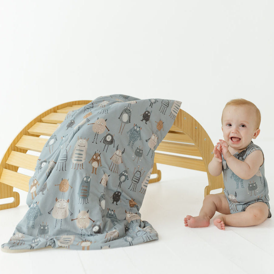 A baby wearing a matching light blue-gray romper sits beside a wooden bench draped with a matching patterned blanket. The fabric features a whimsical print of small cartoon animals in muted brown, beige, and gray tones.