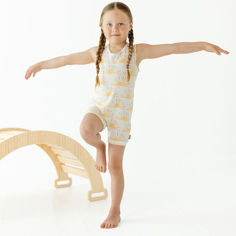 A young girl with braided hair stands barefoot on a white studio background, balancing with outstretched arms and one leg raised. She is wearing a sleeveless white romper with a yellow sunburst print.
