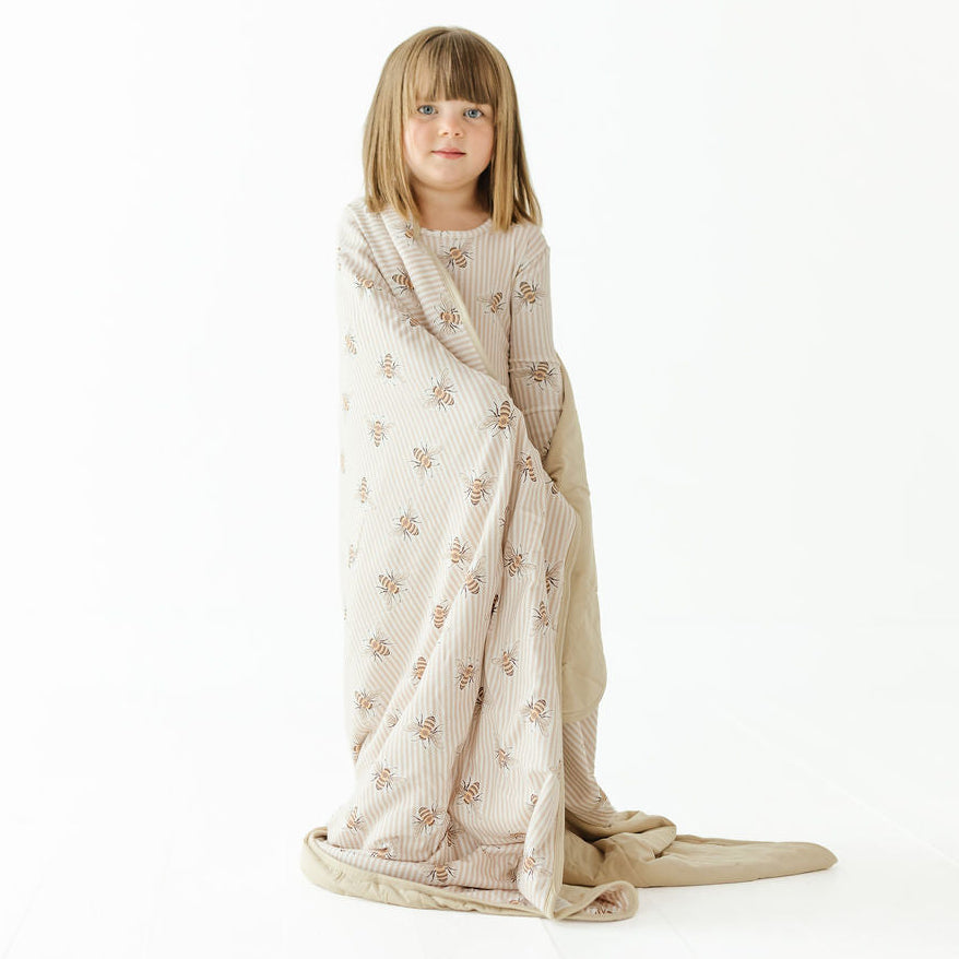 A young girl with blonde hair sits wrapped in a ribbed knit blanket with a small floral pattern, set against a seamless white studio background.
