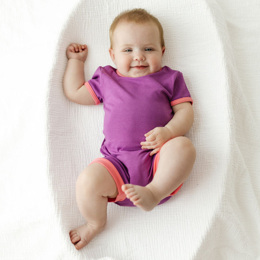 A baby lying on a white textured blanket, wearing a purple short-sleeve romper with pink trim on the sleeves and leg openings.