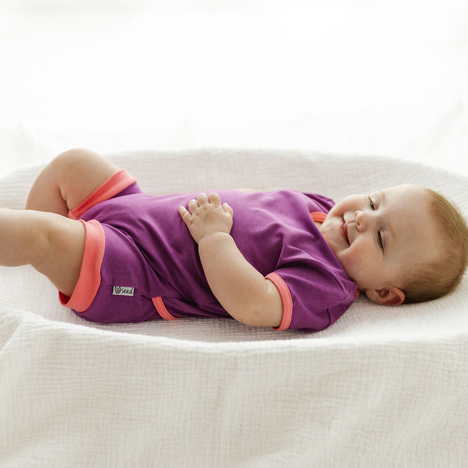 A baby lying on their back on a white textured blanket, wearing a purple short-sleeve romper with coral pink trim.