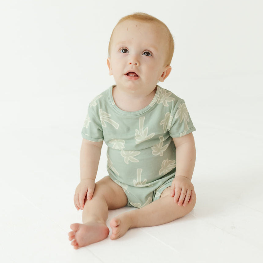 A baby sitting on a white studio floor wearing a sage green short-sleeve bodysuit with a light beige leaf pattern.