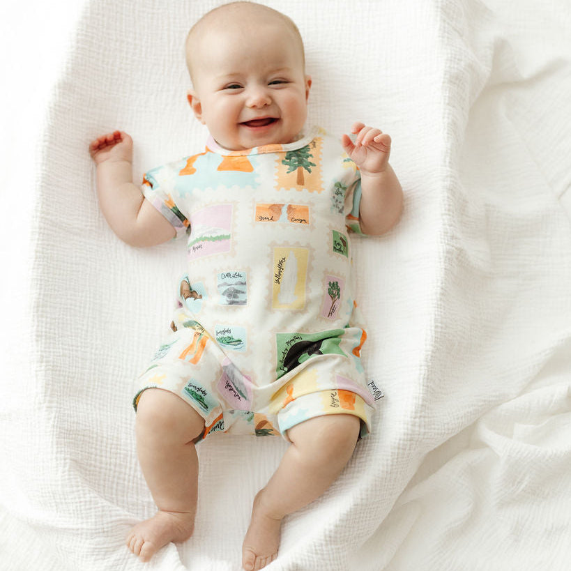 A smiling baby lying on their back on a white textured muslin blanket, wearing a short-sleeve romper with a colorful tropical and landscape print.