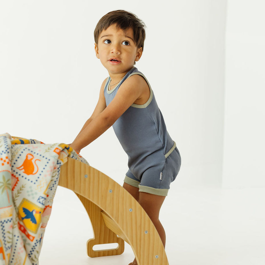 A toddler boy wearing a slate blue sleeveless romper stands on a wooden climbing frame, holding a patterned blanket in a bright, minimalist studio setting.
