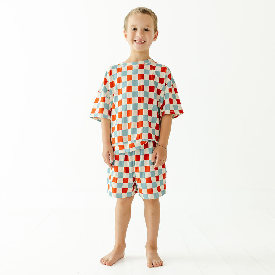 A young boy stands barefoot against a plain white studio background, wearing a matching short-sleeve pajama set with an orange, red, and light blue checkerboard pattern.