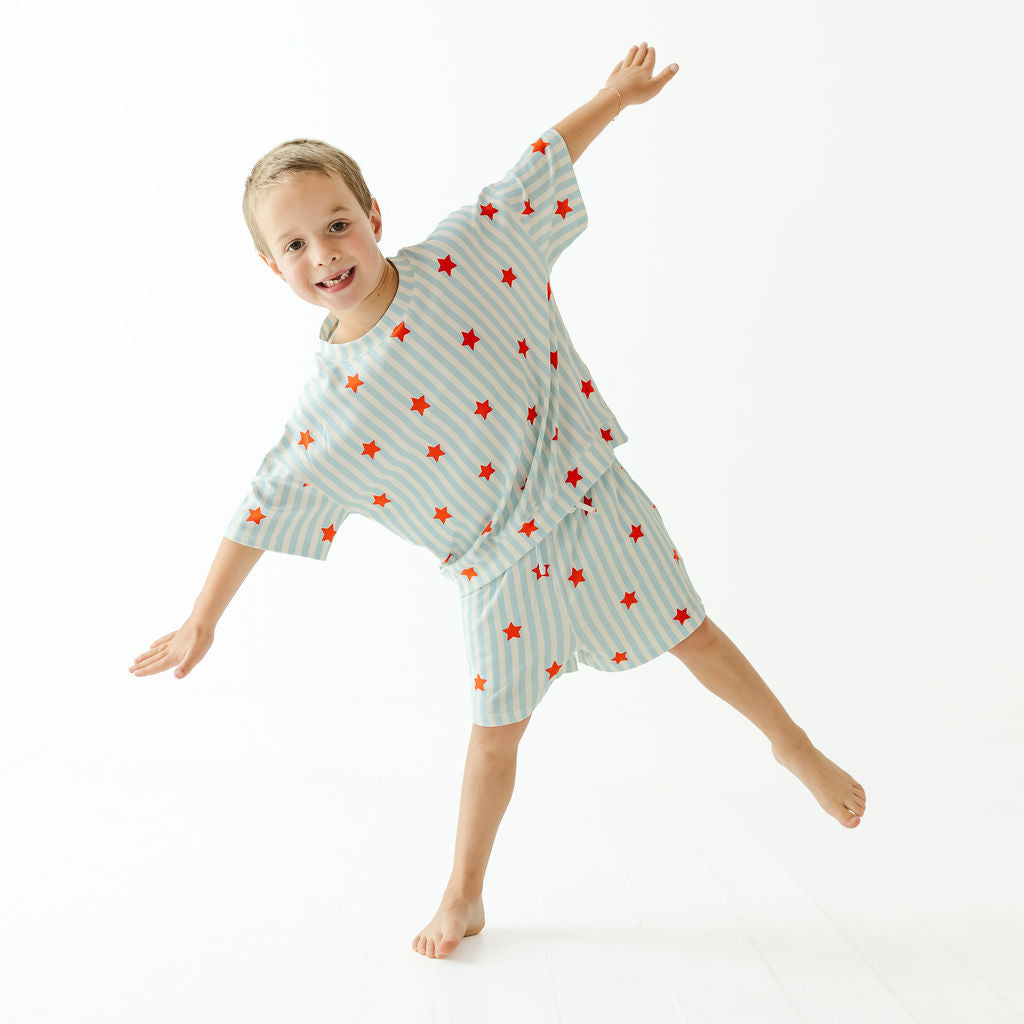 A young boy wearing a light blue and white striped pajama set with red star patterns, posing dynamically on a seamless white studio background.