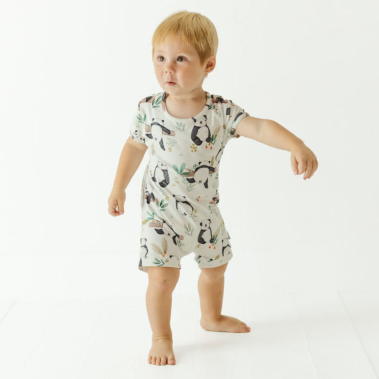 A toddler standing barefoot on a white studio floor, wearing a short-sleeve romper with an allover penguin print on a light cream background.
