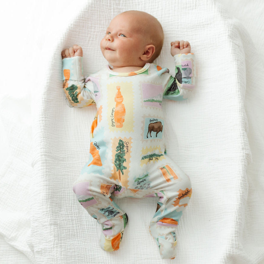 A baby lying on their back on a white textured muslin swaddle blanket, wearing a long-sleeve footed sleeper with a colorful patchwork animal print.
