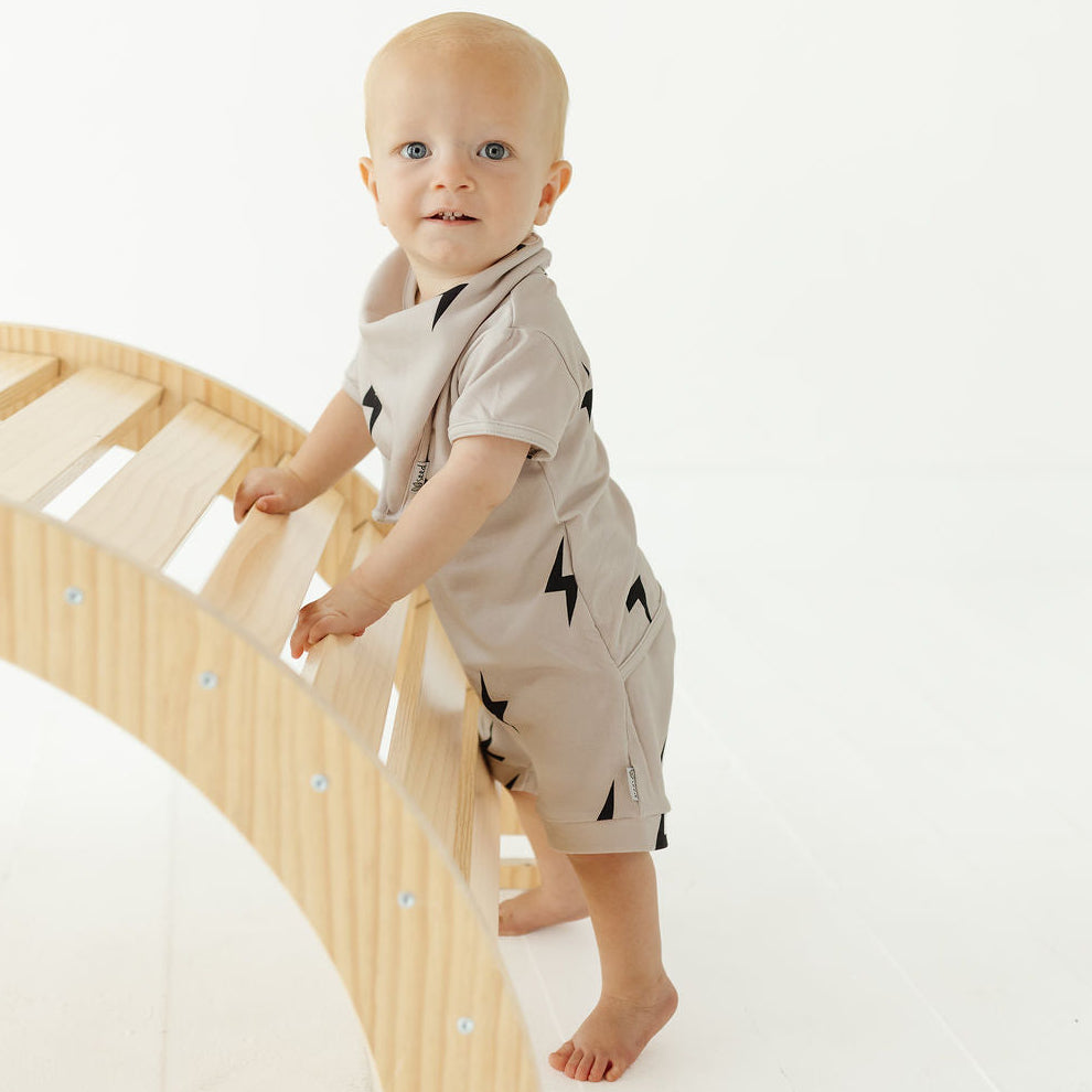 A baby wearing a beige short-sleeve romper with black lightning-bolt prints stands on a curved wooden slide against a clean white background.
