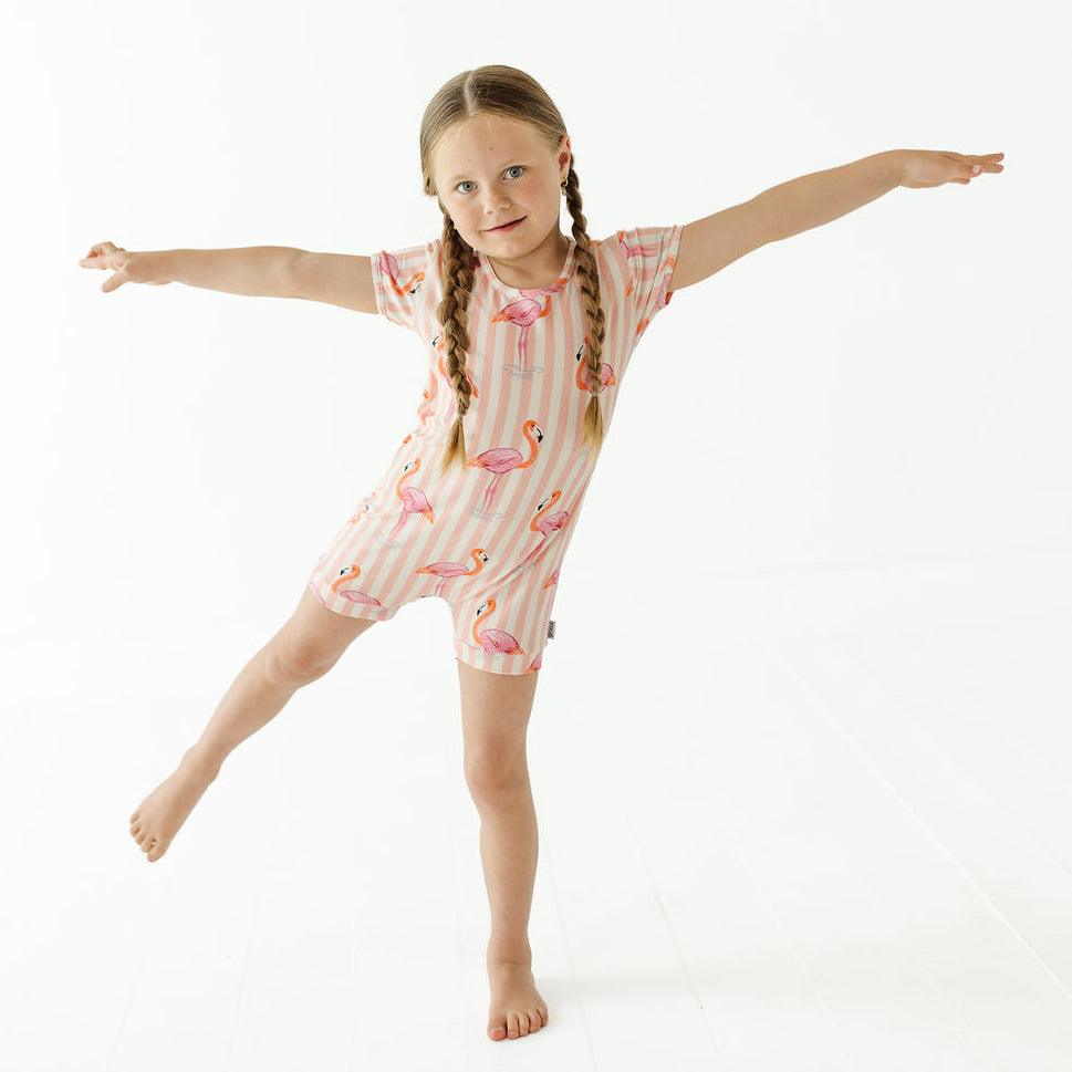A young girl with braided hair poses in a short-sleeve pajama romper with pink flamingo prints and vertical stripes on a plain white studio background.