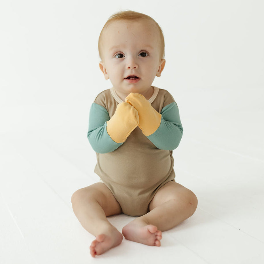 A baby sitting on a white studio floor wearing a tan and teal long-sleeve romper with mustard yellow mittens.