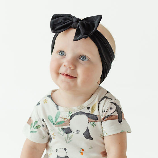 A baby sitting on a white studio floor wearing a short-sleeve cream bodysuit with a panda print and a black velvet bow headband.