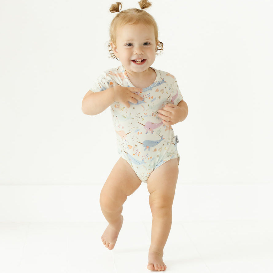 A smiling toddler girl with two small pigtails is wearing a short-sleeve one-piece bodysuit and running barefoot on a seamless white studio background. The bodysuit has a light cream base with a pastel multicolor print of cartoon dinosaurs and stars.