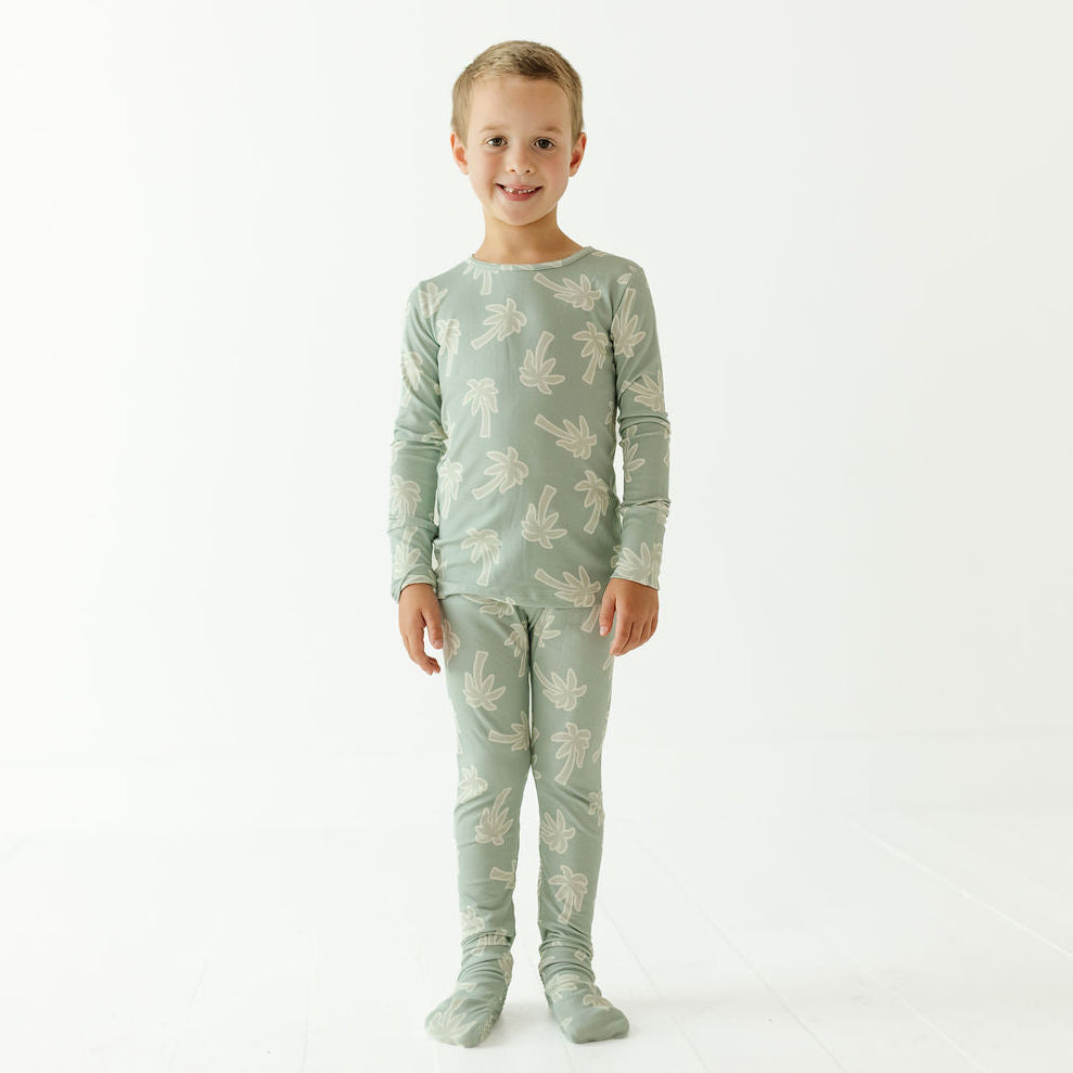 A young child standing against a plain white studio background, wearing a matching long-sleeve top and leggings pajama set in a pale sage green color with a light beige palm tree pattern.