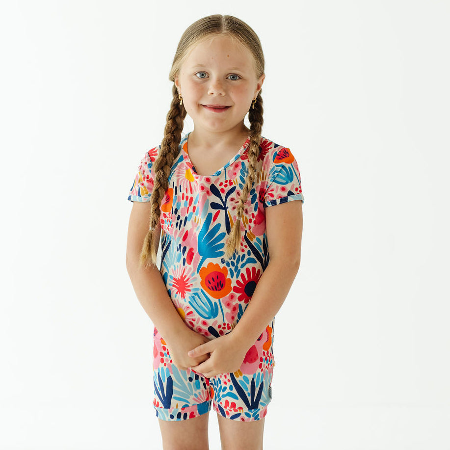 A young girl with braided hair stands against a plain white background wearing a short-sleeve matching set with a colorful floral print.