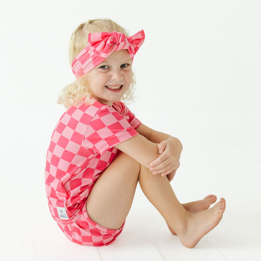 A young girl with blonde hair sits on a white floor, wearing a pink and white checkered short-sleeve romper and a matching checkered bow headband.