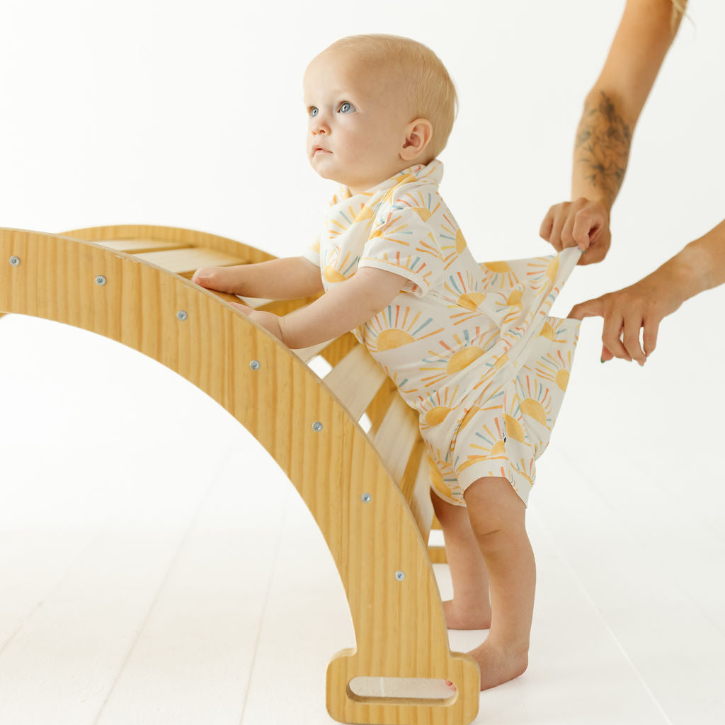 A baby wearing a white and yellow sunburst print romper stands on a wooden climbing ladder, with an adult's hands assisting. The photo is shot in a bright, clean studio setting.