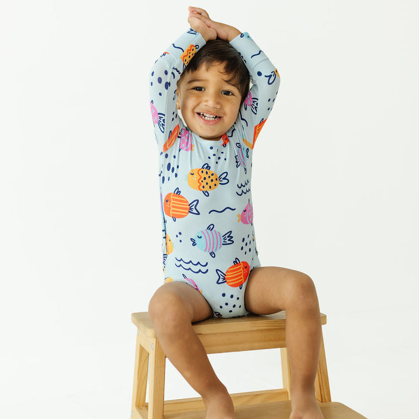 A smiling young child wearing a light blue long-sleeve one-piece swimsuit with a colorful fish pattern, sitting on a small wooden stool against a plain white background.