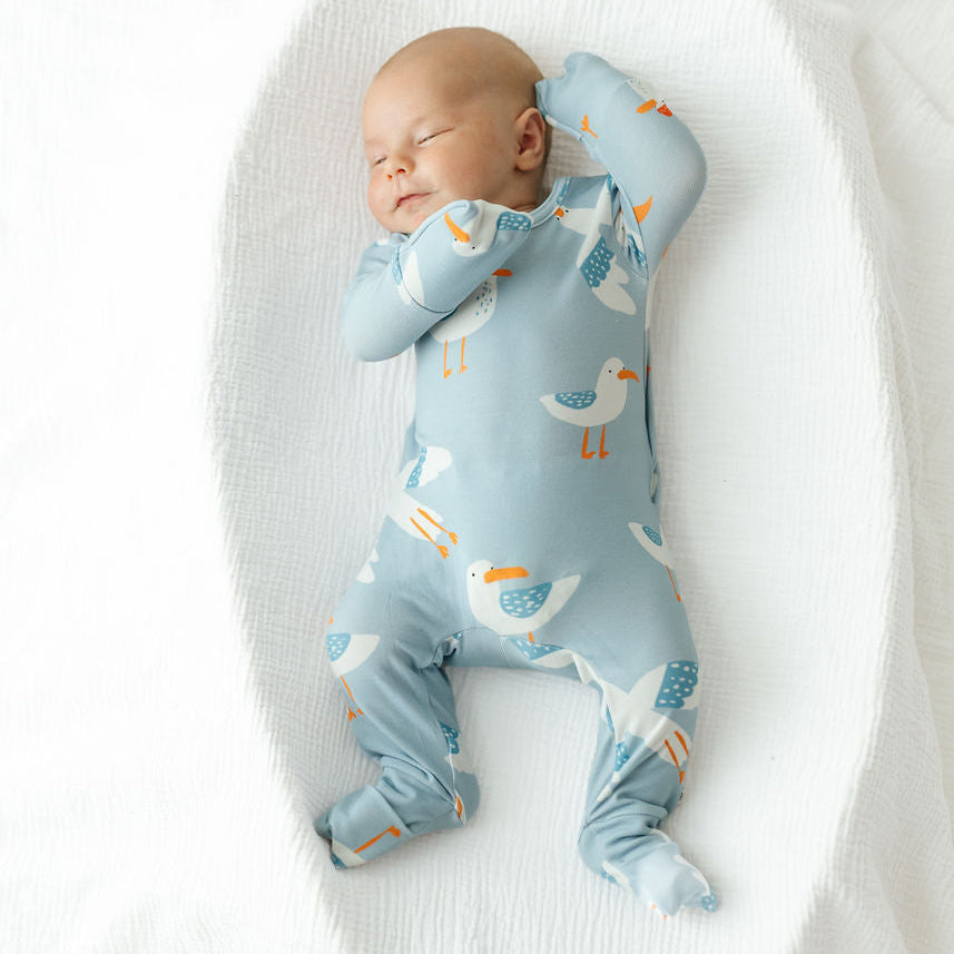 A sleeping newborn baby wearing a light blue footed sleeper with a bird pattern, lying on a white textured blanket.