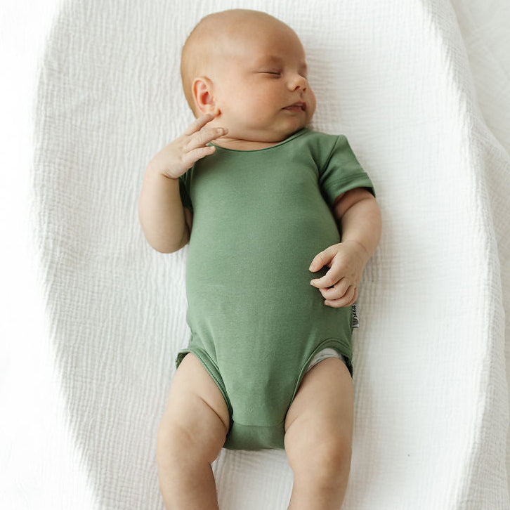 A sleeping newborn baby wearing a sage green short-sleeve bodysuit, lying on their back inside a white textured bassinet or changing pad.