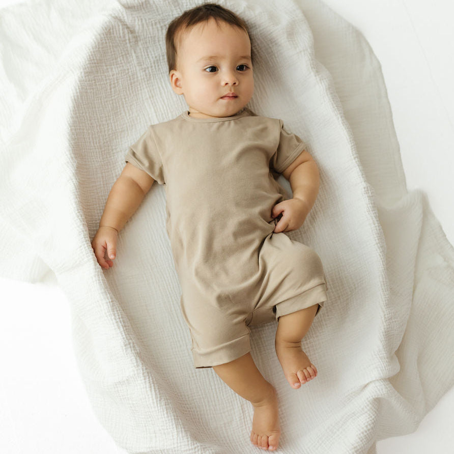 A baby lying on a white textured blanket, wearing a short-sleeve beige romper.