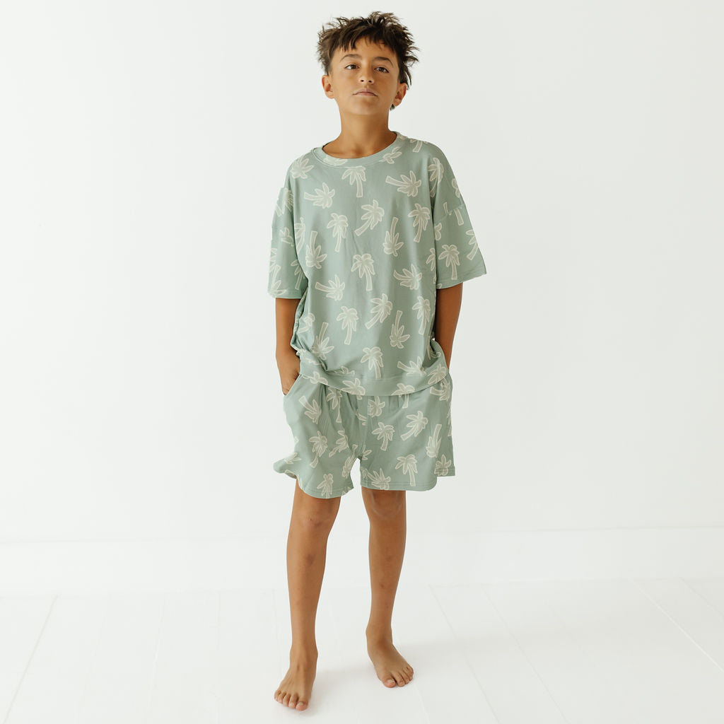 A child model wearing a light sage green two-piece pajama set with a white palm tree pattern, standing against a clean white studio background.