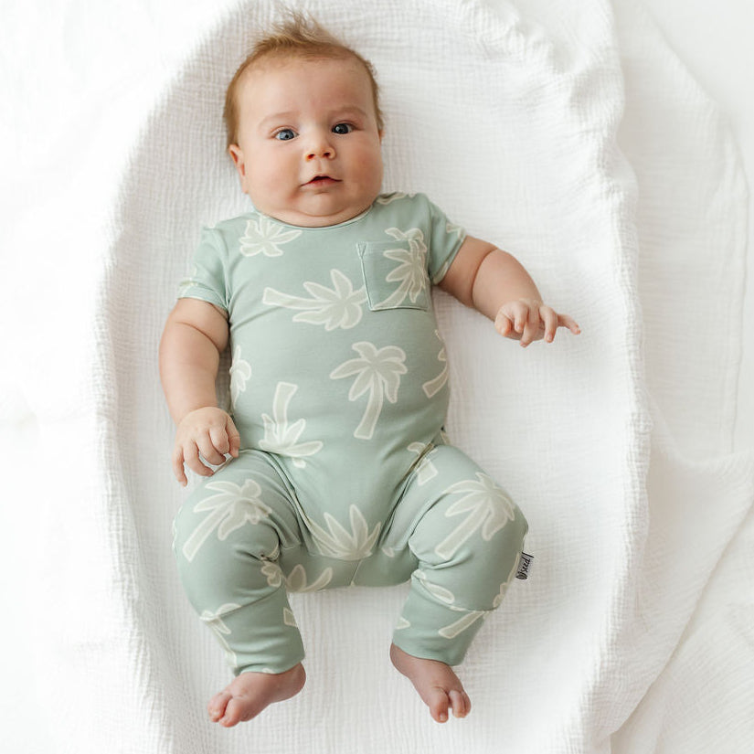 A baby lying on a white textured swaddle blanket, wearing a pale sage green short-sleeve romper with a white palm tree pattern.