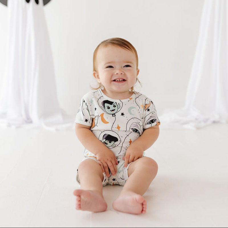 A smiling baby sits barefoot on a white floor, wearing a white short-sleeve romper with a black, gray, and orange space-themed print. In the background, two white ghost decorations with black pointed hats stand on either side.