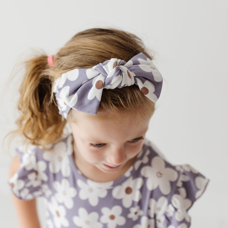 A young girl with blonde hair in a ponytail wearing a lavender and white floral headband with a matching floral top, looking down against a plain white background.