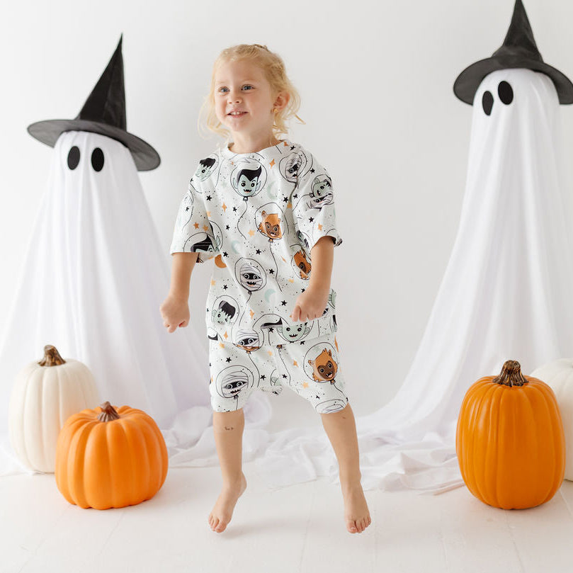 A young child stands barefoot in a studio wearing a white short-sleeve pajama set with a black cat print. The scene is decorated for Halloween with two ghost props wearing black hats, orange and white pumpkins, and a white backdrop.