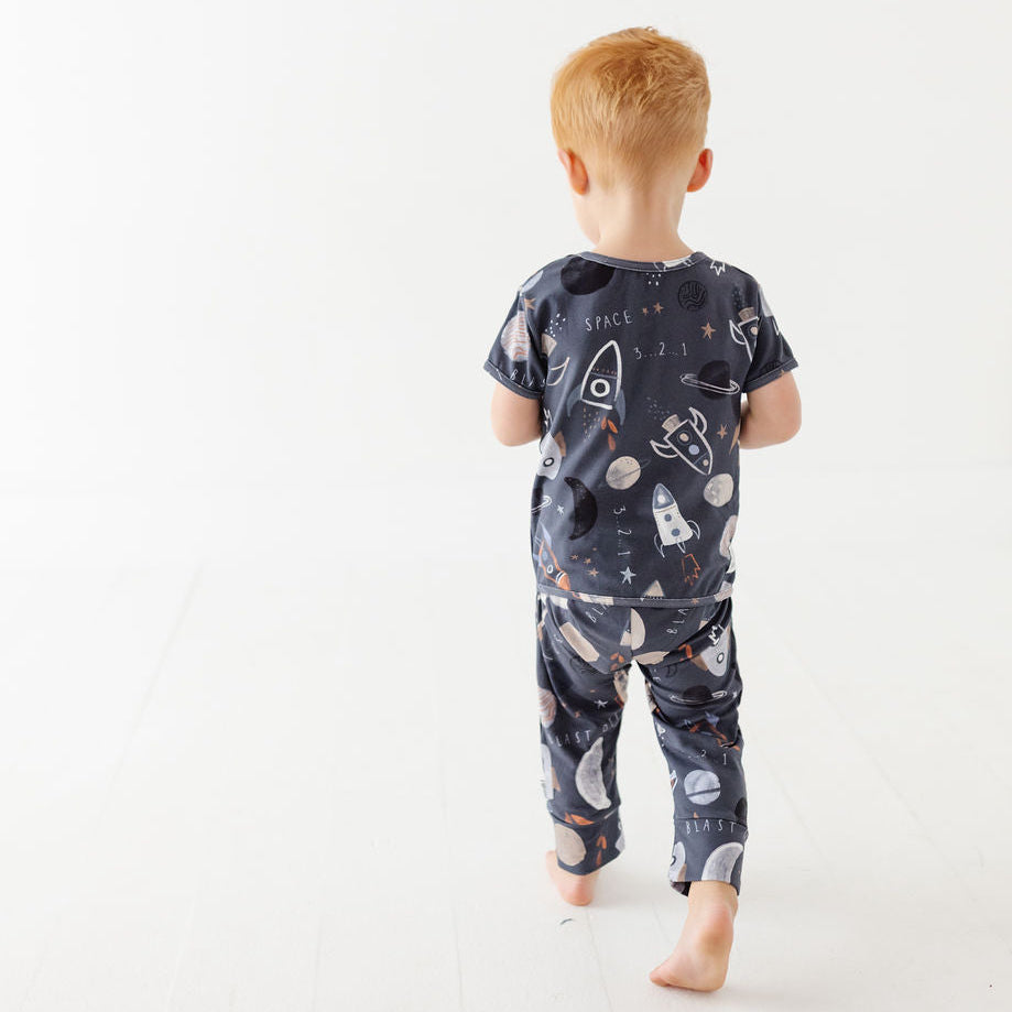 A boy stands on white floor wearing navy romper with space design.