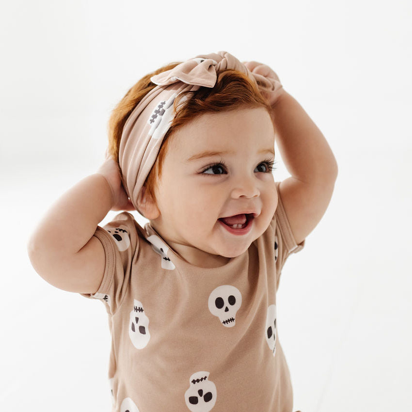 A smiling baby wearing a beige short-sleeve romper with white skull prints and a matching beige bow headband, posed against a bright white background.