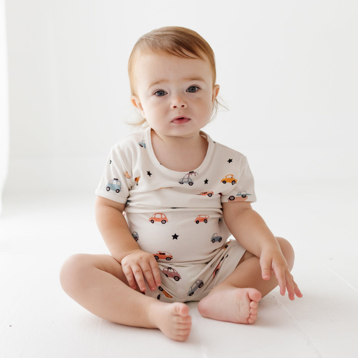 A smiling baby sits barefoot on a white floor, wearing a white short-sleeve romper with a car-themed print.