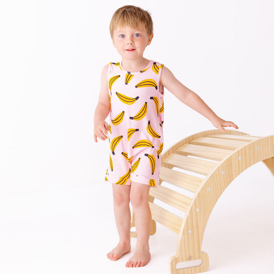Child wearing a pink sleeveless dress with banana prints standing next to a wooden climbing toy on a white background